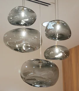 A group of five modern, organic-shaped pendant lights made of smoked translucent glass. The pebble-like globes hang at varying heights, each glowing with a warm internal light against a neutral ceiling and wood-paneled wall.