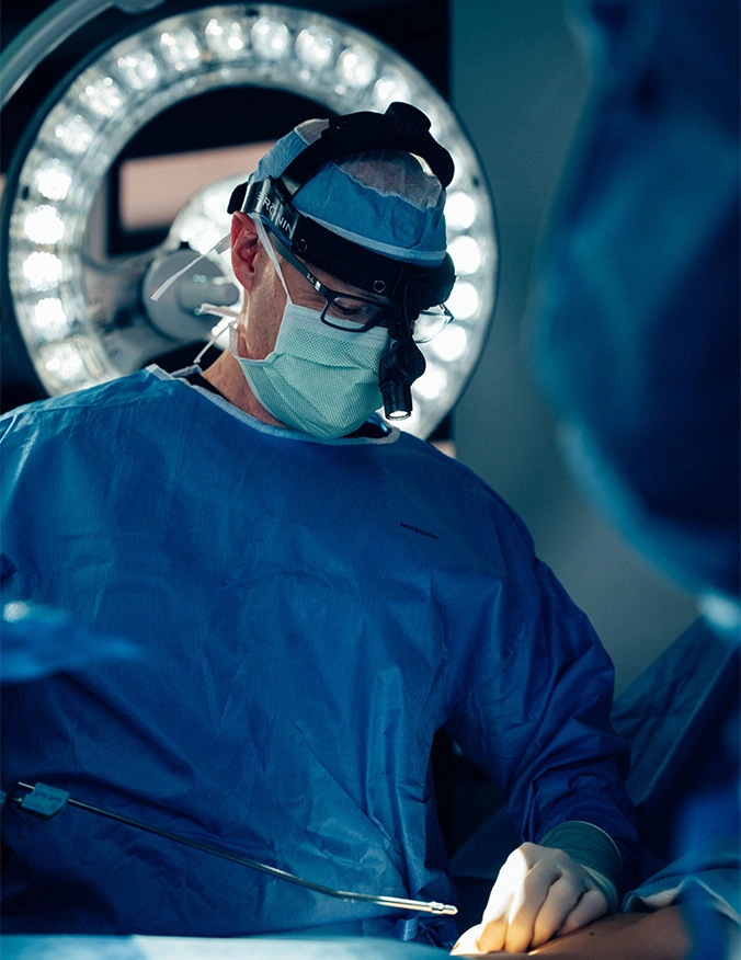 A surgeon in a blue surgical gown, mask, and sterile cap operates under a bright, circular surgical light. He is wearing surgical loupes and glasses, intently focused on the procedure being performed in a sterile operating room environment. - Abdominal etching in Draper, UT
