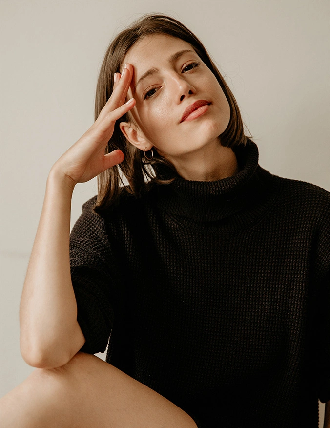 A close-up portrait of a woman with shoulder-length brown hair wearing a black turtleneck sweater. She tilts her head slightly and rests her fingers against her temple, looking directly at the camera with a calm, neutral expression. - Aveli cellulite reduction in Draper, UT