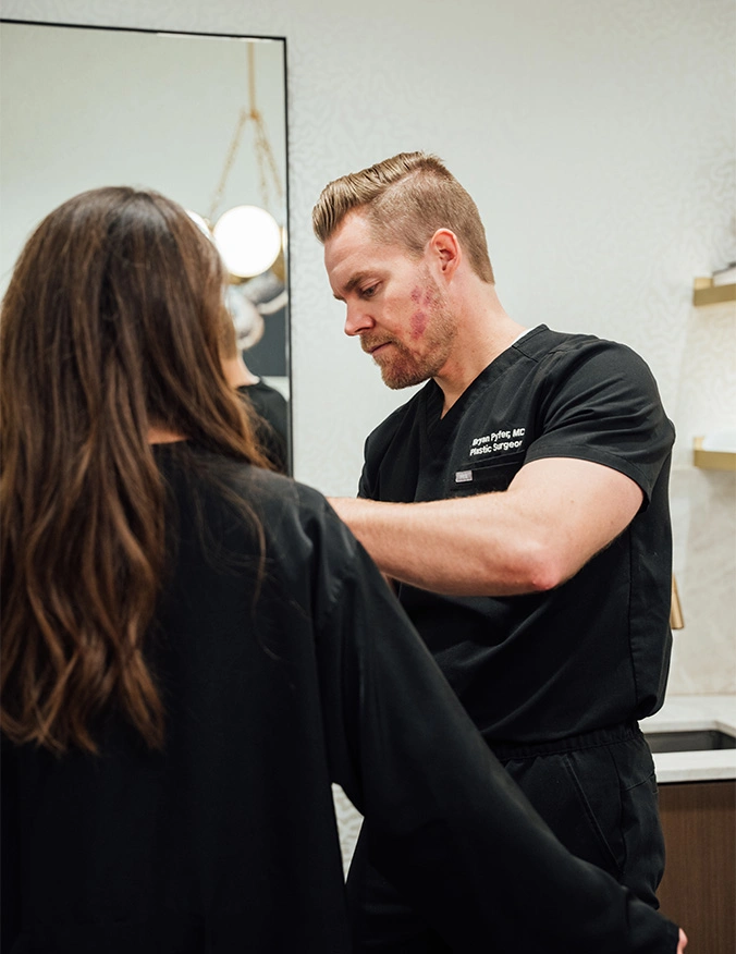 A male plastic surgeon in black scrubs, identified by his name tag as Bryan Pyfer, MD, performs a consultation or procedure on a female patient. The surgeon is seen in profile, focused on his work, while the patient stands with her back to the camera in a black medical gown. - Breast augmentation in Draper, UT