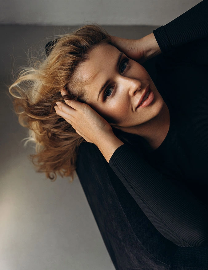 A high-angle portrait of a woman with blonde hair, wearing a black top. She has her hands tucked into her hair near her temples, gently pulling back to highlight a smooth forehead and lifted brow area while looking directly at the camera. - Brow lift in Draper, UT