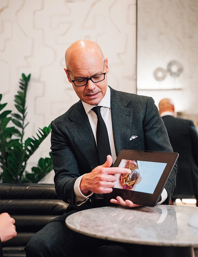 A doctor wearing a dark grey suit, white shirt, and black tie, sits and points to a medical image displayed on a tablet. He is wearing glasses and appears to be explaining the image to someone off-camera in a professional, modern office setting. - Capsulectomy in Draper, UT