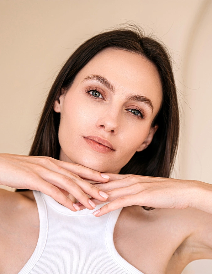 A head-on portrait of a woman with dark hair and blue eyes, looking calmly at the camera. She is resting her chin on the backs of her hands, which are overlapped in front of her neck. She is wearing a white ribbed tank top against a neutral, warm-toned background. - Chemical peels in Draper, UT