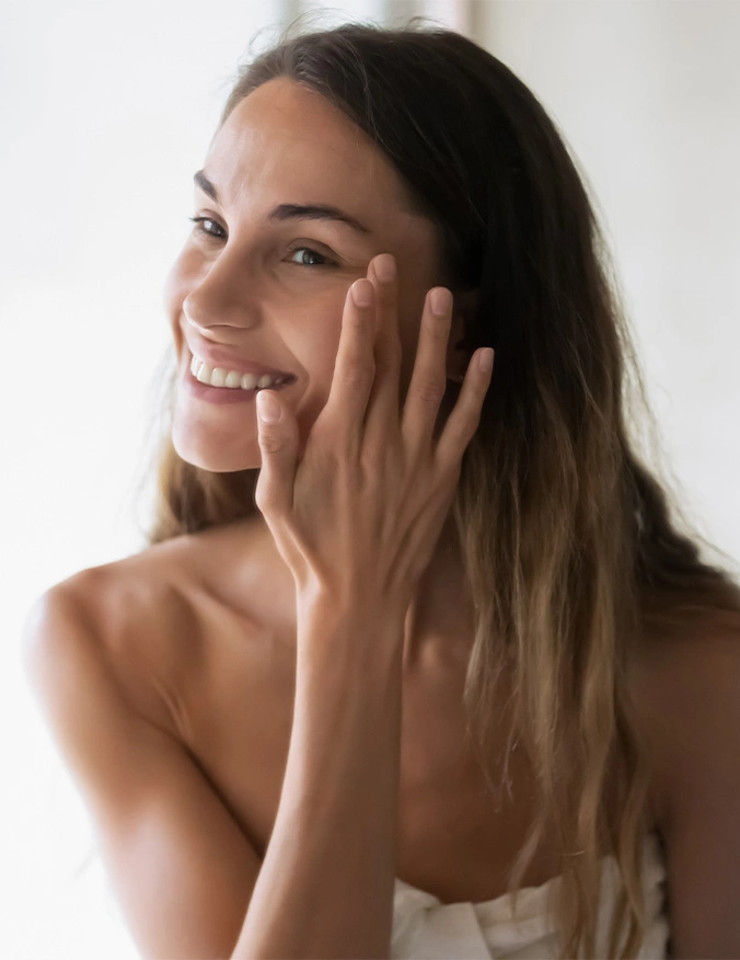 A close-up shot of a smiling woman with long brown hair, looking directly toward the camera. She is touching her cheek with her fingertips, highlighting her smooth and bright skin. The lighting is high-key and airy, creating a fresh and vibrant aesthetic. - DiamondGlow in Draper, UT