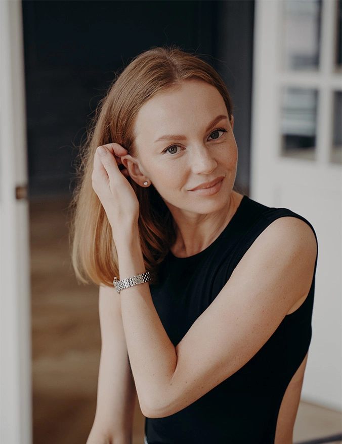 A woman with reddish-brown hair and light freckles smiles warmly at the camera. She is tucked into a black sleeveless top and lightly brushes a hand behind her ear, showing off her rejuvenated facial contours in a brightly lit indoor setting. - Facial fat grafting in Draper, UT