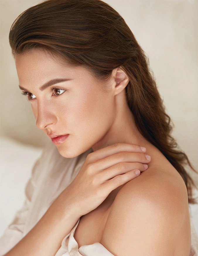 A profile portrait of a woman with long brunette hair looking pensively to the side. She has a clear, radiant complexion and is resting her hand gently on her shoulder. The background is soft and out of focus, emphasizing her refined features and glowing skin. - Facials in Draper, UT