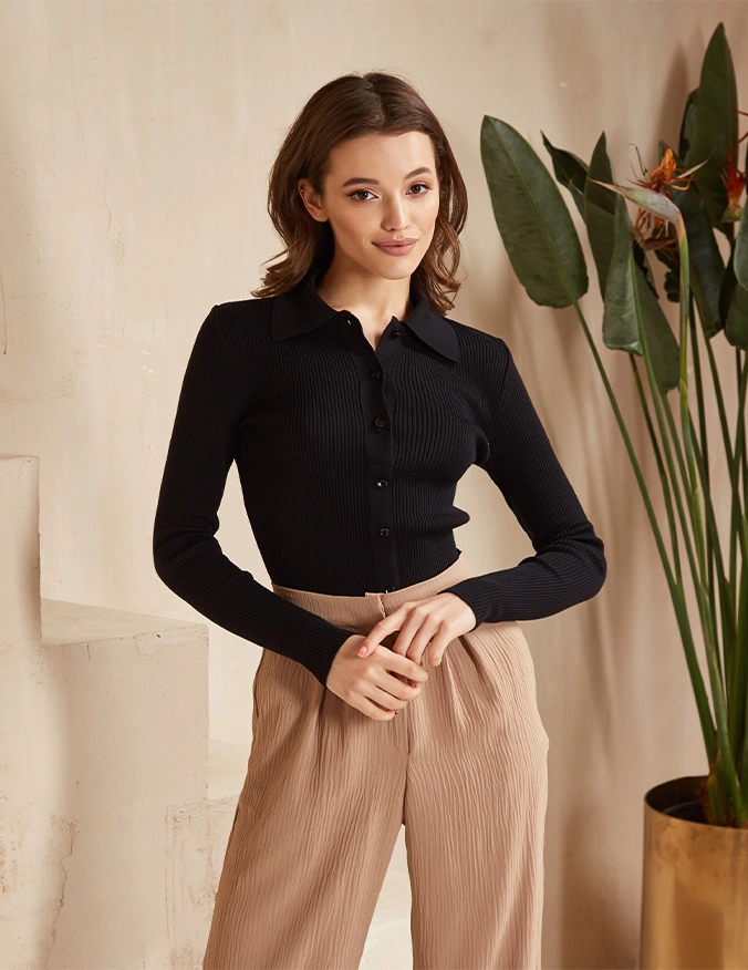 A woman with long brown hair standing indoors, wearing a black ribbed long-sleeve polo shirt and pleated tan trousers, posed with her hands clasped in front. - Food sensitivity testing in Draper, UT