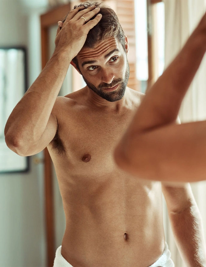 A shirtless man with a beard and dark hair looks into a bathroom mirror, running one hand through his hair. He is wearing a white towel around his waist, and the reflection shows his side profile as he inspects his chest and torso in the bright, natural morning light. - Gynecomastia in Draper, UT
