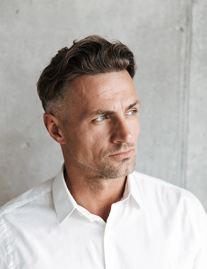 A professional headshot of a middle-aged man with salt-and-pepper hair and blue eyes. He is wearing a crisp white button-down shirt and looking to the side with a calm, confident expression. The background is a neutral, textured gray concrete wall. - Hair transplant in Draper, UT