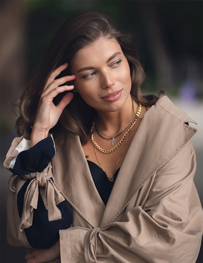 A woman with brown hair and glowing skin looks off-camera with a slight smile. She is wearing a tan trench coat draped over her shoulders, a black top, and layered gold chain necklaces. One hand is raised to her hair, and her coat sleeves feature distinctive bow-tie details at the wrists. - Halo Tribrid in Draper, UT