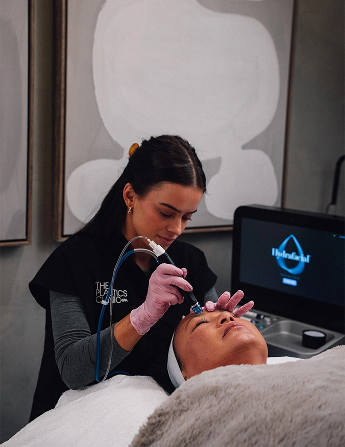 A clinical aesthetician wearing pink gloves and a black "The Plastics Clinic" uniform performs a facial treatment on a patient. The patient is lying down with a white headband, while the specialist uses a handheld HydraFacial device on her forehead. A monitor displaying the HydraFacial logo is visible in the background. - hydrafacial in Draper, UT