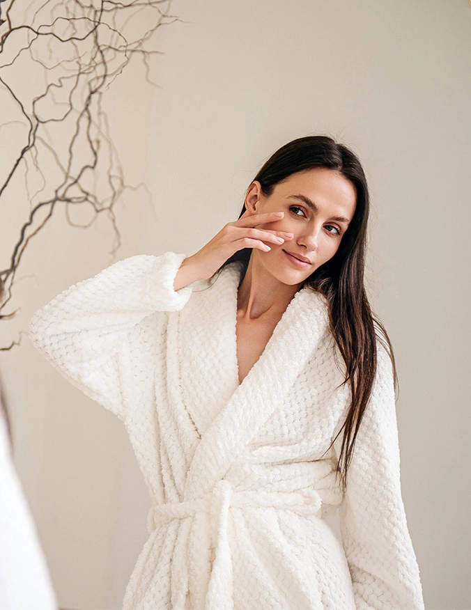 A woman with long dark hair stands in a bright, airy room wearing a plush white bathrobe. She gently touches her cheek with her fingertips, smiling softly at the camera to highlight her smooth, glowing skin. - Laser skin rejuvenation in Draper, UT