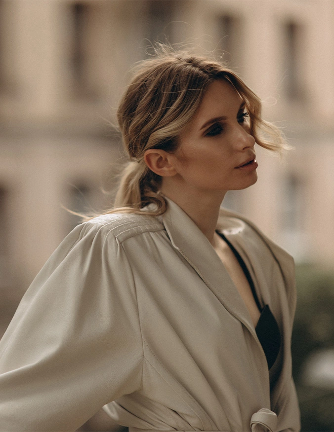 A cinematic portrait of a blonde woman looking toward the side against a blurred urban background. She is wearing a beige, structured wrap coat with a black top underneath. The lighting is soft and natural, emphasizing her clear complexion and refined facial features. - Laser skin resurfacing in Draper, UT