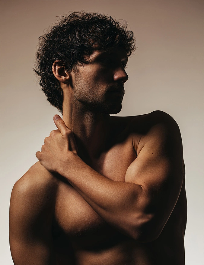 A profile shot of a muscular man with dark, curly hair and light stubble. The lighting is moody and atmospheric, silhouetting his face while highlighting the contours of his neck, shoulder, and bicep. He is shirtless, with one hand resting on his opposite shoulder against a neutral, warm-toned background. - Male liposuction in Draper, UT