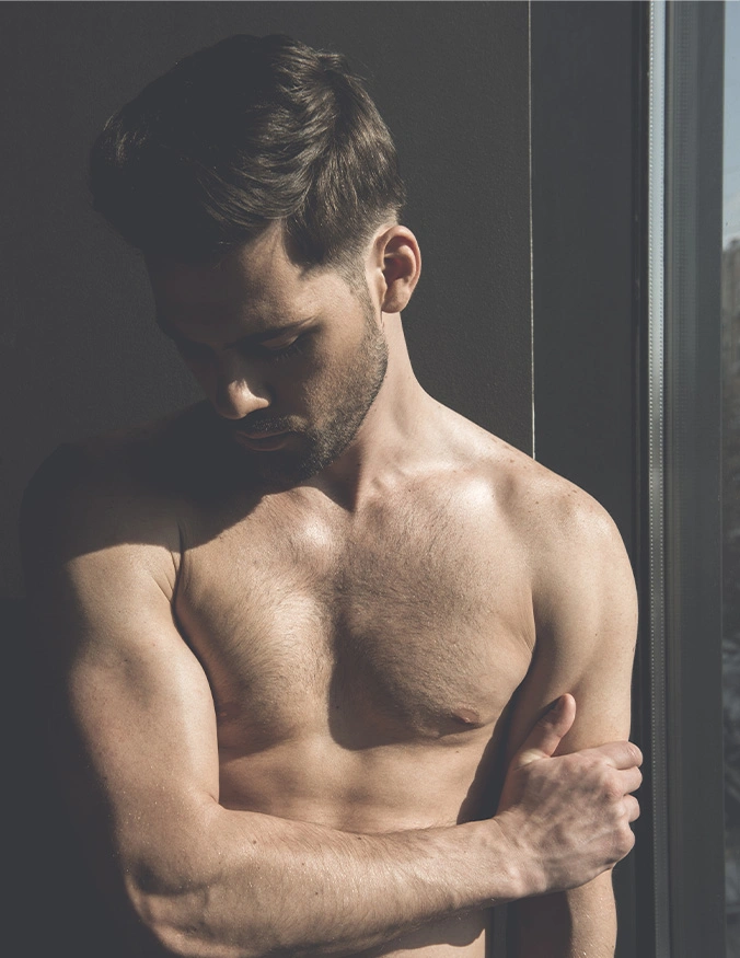 A dramatic, high-contrast photograph of a shirtless man standing by a window. His head is tilted down, casting a shadow over his face, while bright sunlight highlights the definition of his chest and muscular arms. He has dark, styled hair and a neatly trimmed beard, with his hand resting on his forearm in a reflective pose. - Male massive weight loss in Draper, UT