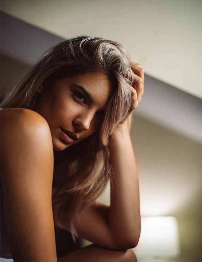 A vertical, close-up portrait of a woman with blonde-highlighted hair, looking directly at the camera with an intense, serene gaze. She is resting her head on her hand, with her fingers tucked into her hair. The lighting is soft and warm, emphasizing her clear skin and the natural texture of her hair. - RF microneedling in Draper, UT