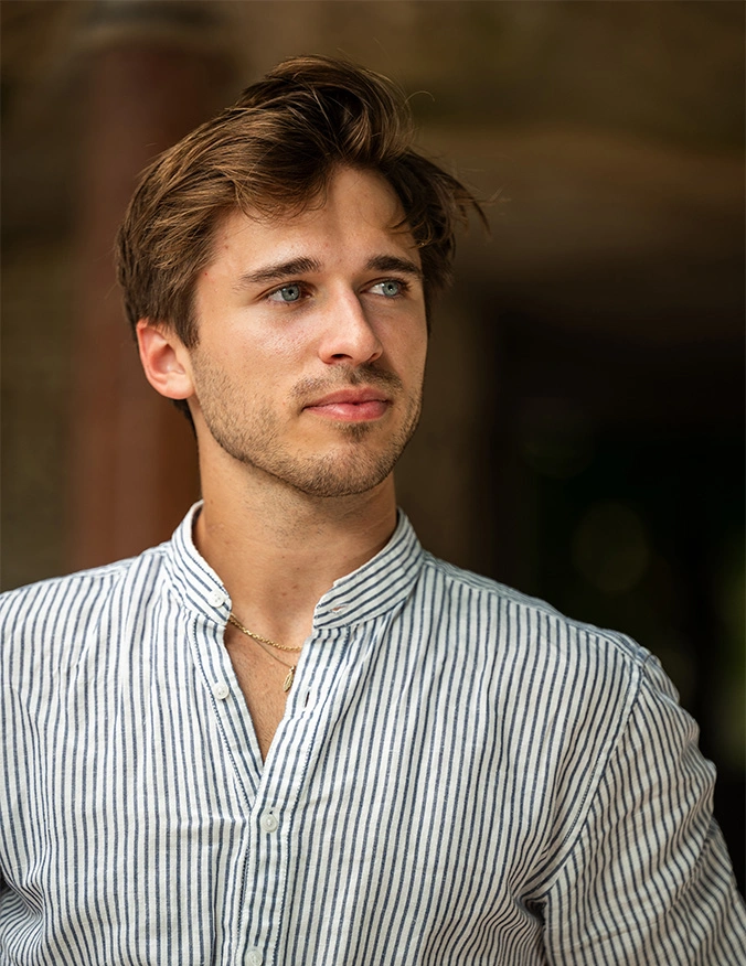 A portrait of a young man with light brown hair and blue eyes, wearing a blue and white striped button-down shirt. He is looking off-camera to the right with a faint, confident smile against a blurred, warm-toned architectural background. - Septoplasty in Draper, UT