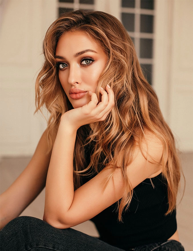 A woman with voluminous, wavy honey-blonde hair and dramatic eye makeup poses with her chin resting on her hand. She is wearing a black sleeveless top and dark jeans, looking intensely at the camera in a warm, elegantly lit indoor setting. - Skinvive in Draper, UT