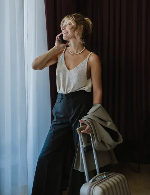 A woman stands near a window with sheer white curtains, smiling while talking on a mobile phone. She is dressed in a light-colored knit tank top and dark wide-leg trousers, holding a tan blazer in one hand and a striped rolling suitcase in the other. - in Draper, UT