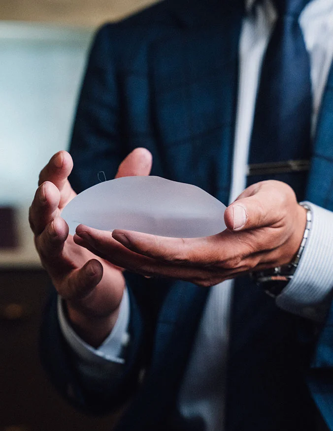 A medical professional in a blue suit holds a round, translucent silicone breast implant while consulting with a patient, whose shoulder and back are visible in the foreground. - Breast augmentation in Draper, UT