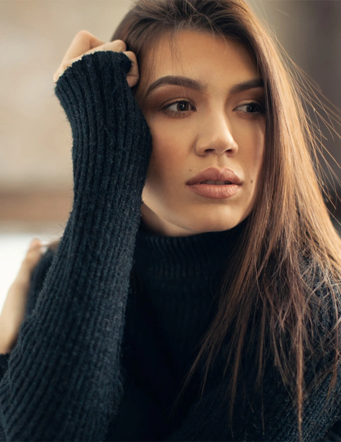 A focused portrait of a woman with long brown hair, wearing a dark knit turtleneck. She looks away from the lens, highlighting the clarity and openness of her eye area and the natural symmetry of her features. - Eyelid surgery in Draper, UT