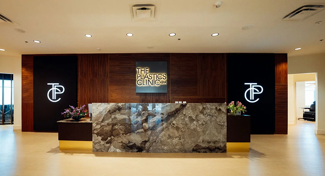 The reception area of "The Plastics Clinic + Spa," featuring a large grey marble desk against a dark wood-paneled wall. Two glowing "TP" logos flank the central backlit clinic sign.