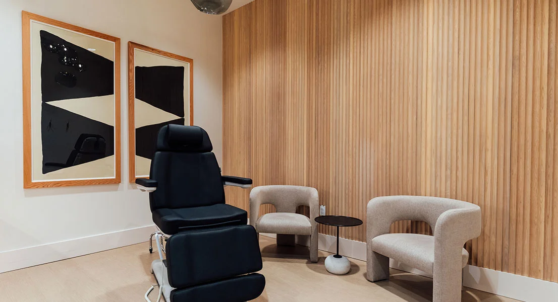 A modern medical treatment room featuring a black adjustable procedure chair, two beige curved armchairs, and a small side table. The back wall is decorated with vertical wood slats and two large framed abstract prints.