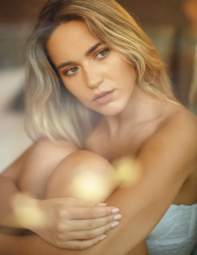 A soft-focus, ethereal portrait of a young woman with blonde hair and green eyes. She is sitting with her knees pulled toward her chest, looking thoughtfully off-camera. The lighting is warm and hazy, with glowing lens flares overlaying the image to create a dreamlike, luminous skin effect. - Microneedling in Draper, UT