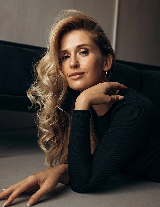 A vertical portrait of a woman with voluminous blonde curls, lying on her side and looking into the camera with a subtle smile. She is wearing a black ribbed long-sleeved top and a small gold hoop earring. The lighting is dramatic and warm, highlighting the contours of her face and the shine in her hair. - moxi in Draper, UT