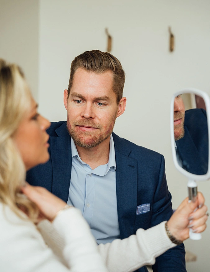 Dr. Bryan Pyfer, wearing a blue blazer and light blue shirt, conducts a consultation with a female patient. The patient holds a white hand mirror, examining her neck and jawline while the doctor looks on with a focused and professional expression. - Neck lift in Draper, UT