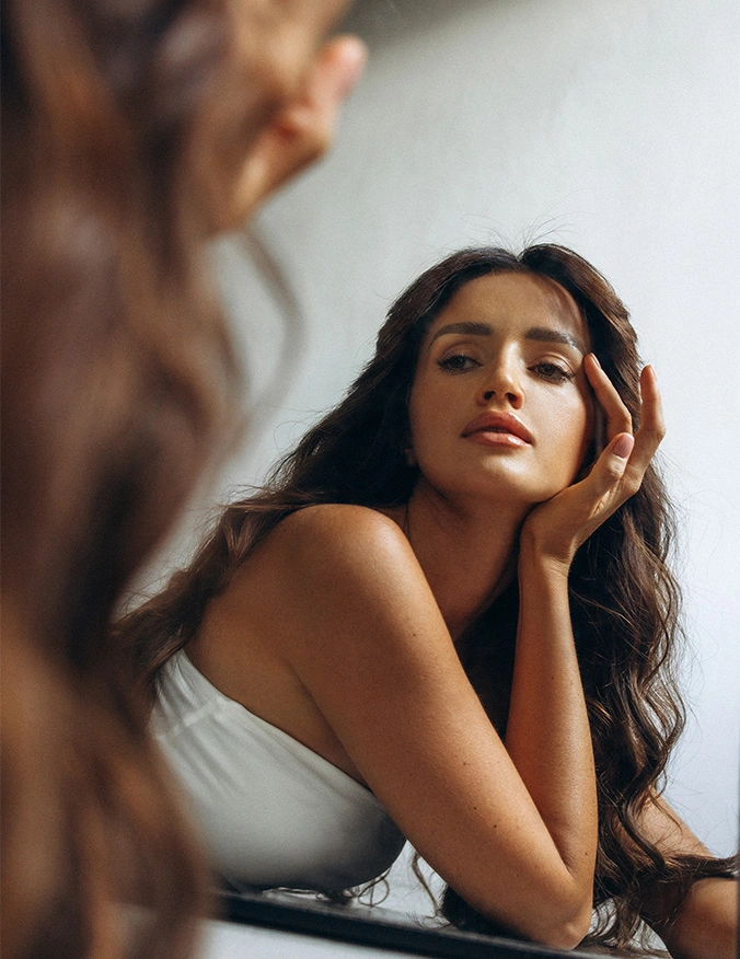 A young woman with long, wavy brown hair gazes thoughtfully into a mirror, resting her chin on her hand. The reflection highlights her clear complexion and defined jawline as she leans forward in a white strapless top against a neutral background. - Neck and chin liposuction in Draper, UT