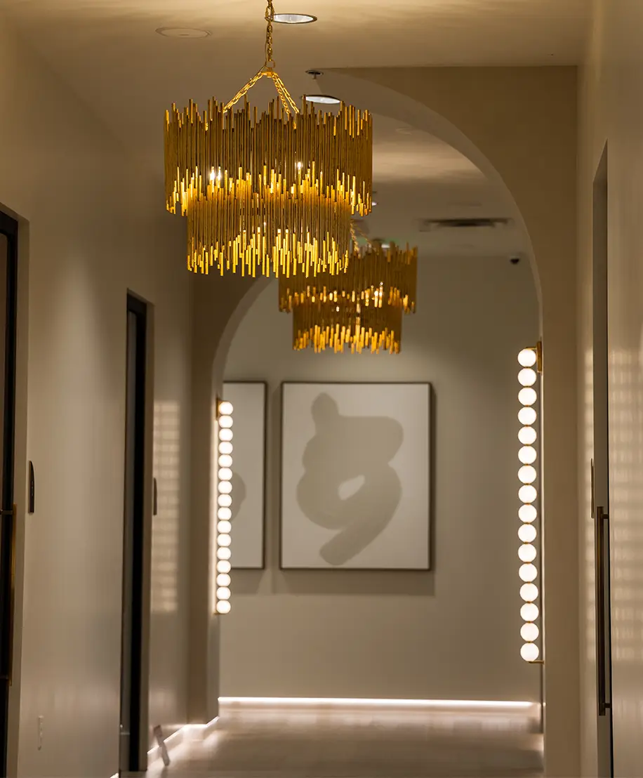 An interior view of a dimly lit, upscale hallway featuring multiple gold-tiered chandeliers hanging from the ceiling and circular glowing wall sconces. Abstract artwork is visible on the walls at the end of the corridor.