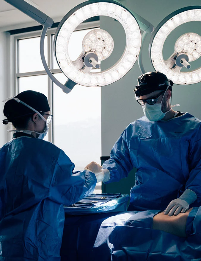 An action shot inside a brightly lit surgical suite. Two medical professionals in blue scrubs, masks, and caps work under large, circular surgical lights. One surgeon wears magnifying loupes while they focused on a procedure. - Tummy tuck in Draper, UT